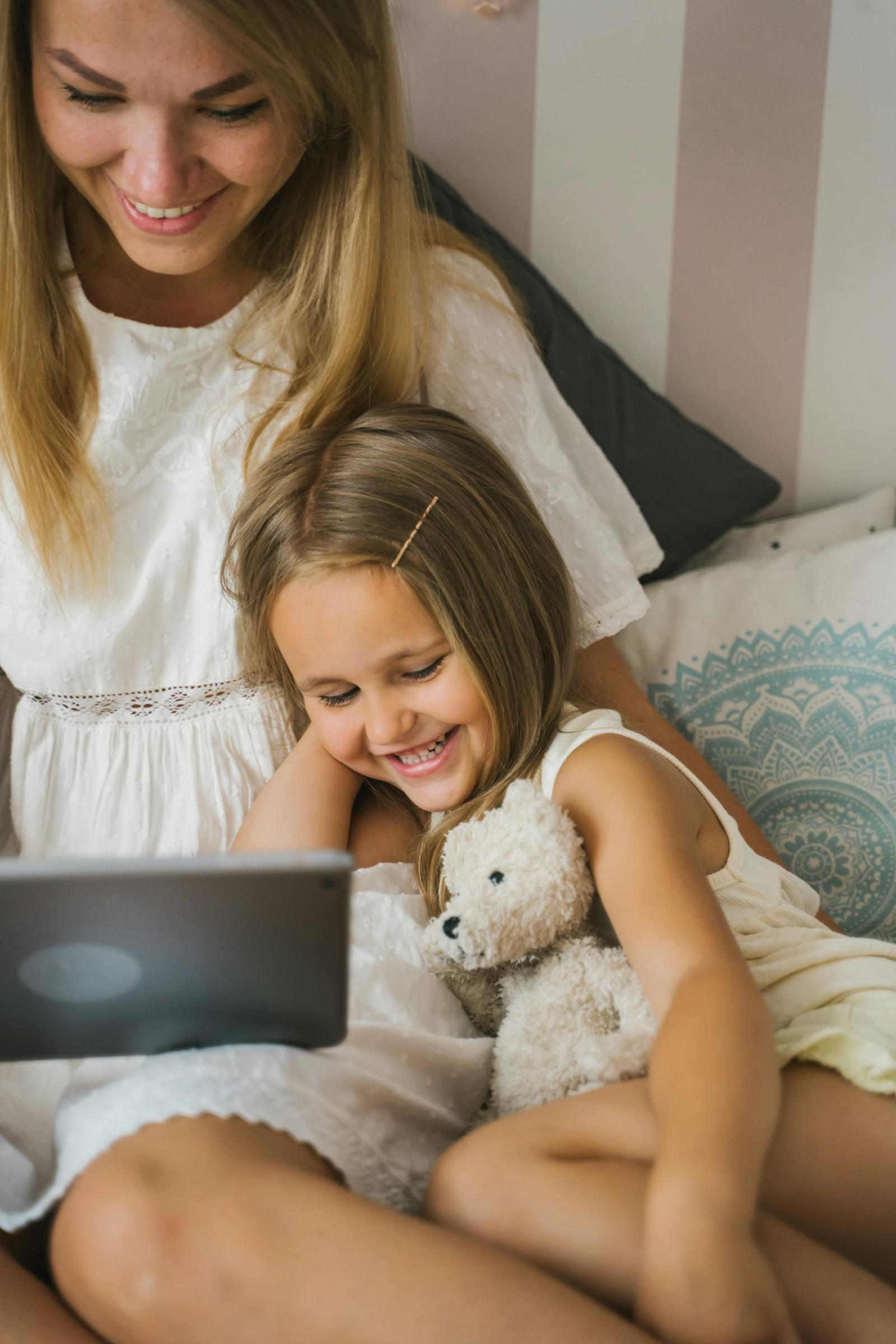 Eine Mutter sitzt mit ihrer Tochter (etwa 5 Jahre) auf dem Bett. Beide schauen gemeinsam auf ein Tablet, lachen. Co-Use-Szene, freudvoll, intim.