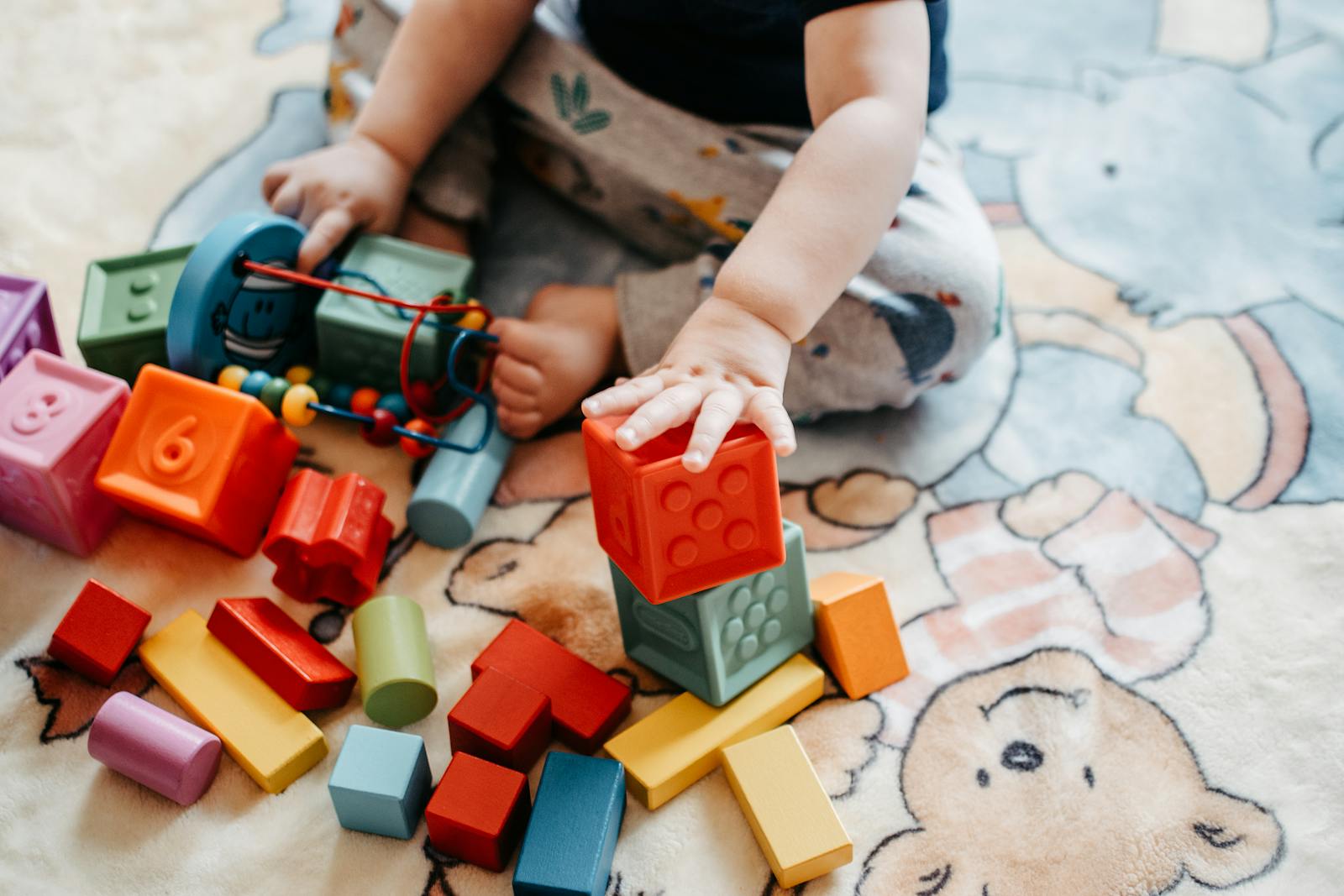 Ein kleines Kind sitzt auf einem Winnie-Puuh-Teppich und stapelt bunte Bauklötze. Ruhige, fokussierte Spielsituation ohne Bildschirm.
