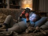 Kleinkind im Schlafanzug auf dem Sofa, Smartphone-Display leuchtet kaltes Blau auf das Gesicht, Stofftier liegt ignoriert daneben.